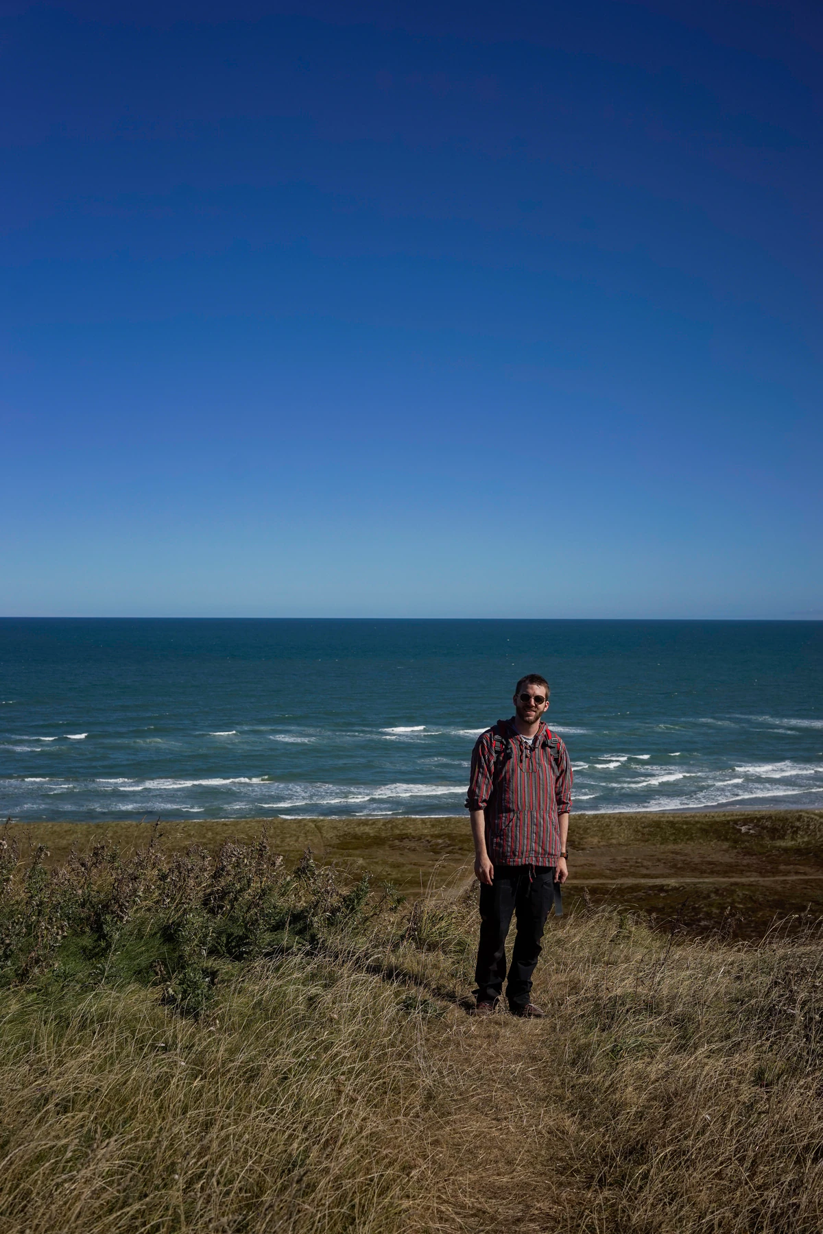 Martin standing in dune grass on a clifftop in Denmark, the sea stretching to the horizon behind him under a cloudless blue sky.
